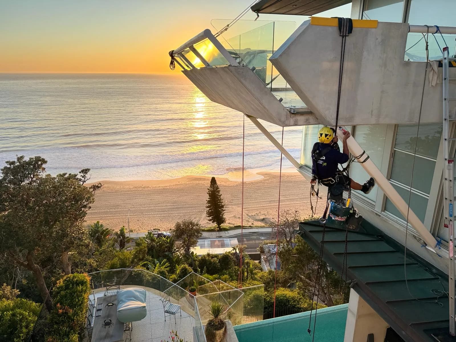 Strata Remedial Services technician on ropes repairing a cliff-top apartment balcony at sunrise
